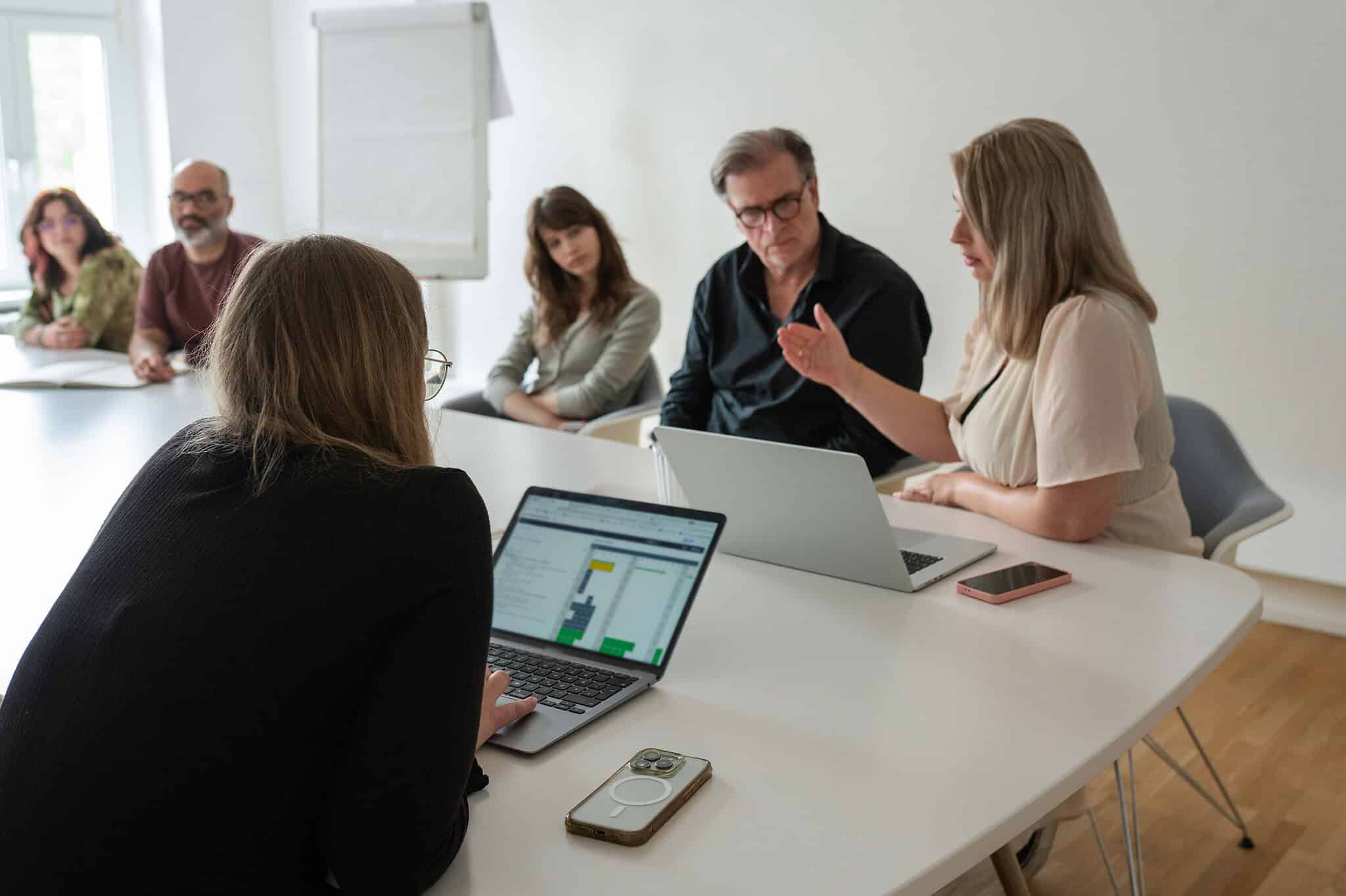 Teammeeting in einem hellen Besprechungsraum mit mehreren Mitarbeitenden, Laptops und Smartphones am Tisch.