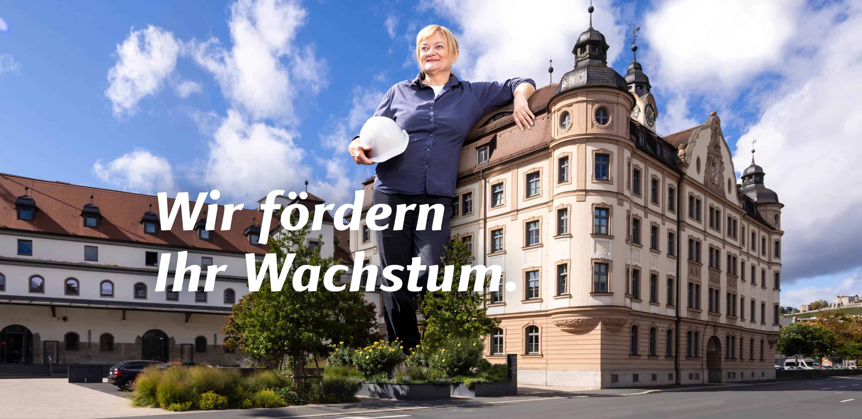 Mitarbeiterin mit Schutzhelm vor einem historischen Gebäude der Stadt Würzburg mit dem Motiv „Wir fördern Ihr Wachstum“.